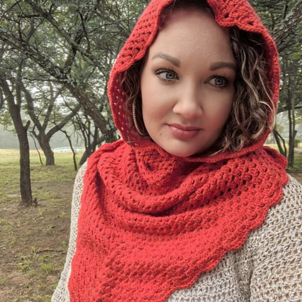 Woman with curly hair looking at the camera, wearing the red crocheted Rooikappie hood and cowl over a cream knit top, with trees and grass behind her.
