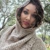Woman looking at the camera with the hood of a crocheted Rooikappie in soft taupe pulled up over her curly hair, with trees behind her.
