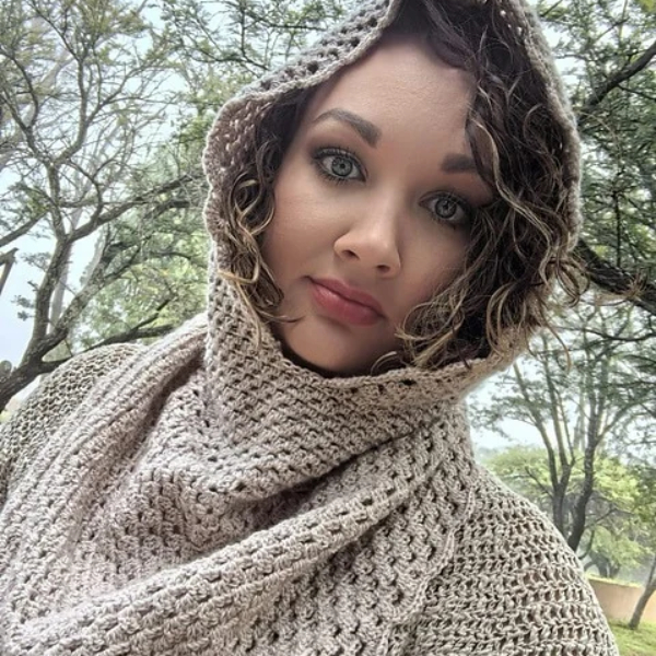 Woman looking at the camera with the hood of a crocheted Rooikappie in soft taupe pulled up over her curly hair, with trees behind her.