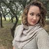 Woman with curly hair looking to the side, wearing the taupe crocheted cowl draped over her shoulders, with a scalloped edge and trees in the background.