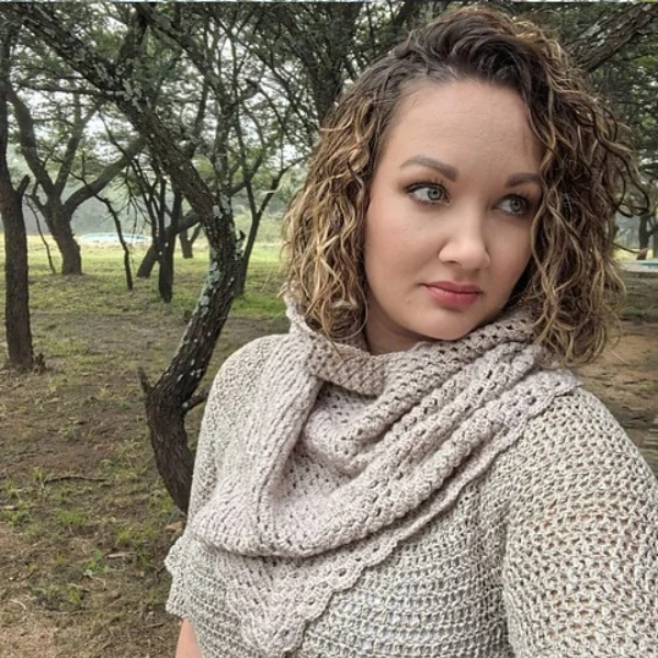 Woman with curly hair looking to the side, wearing the taupe crocheted cowl draped over her shoulders, with a scalloped edge and trees in the background.