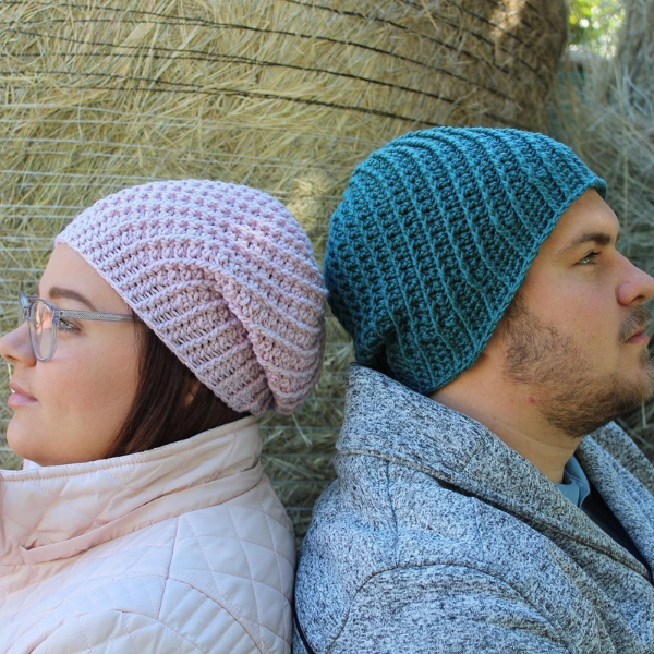 A woman and a man sitting back to back in profile against a hay bale, wearing slouchy crocheted Spiral Beanies, hers in soft pink and his in deep teal.