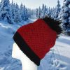Side view of a hand knitted red hat with a wide black ribbed brim and a black pompom, displayed on a white mannequin head in front of snow covered pine trees.