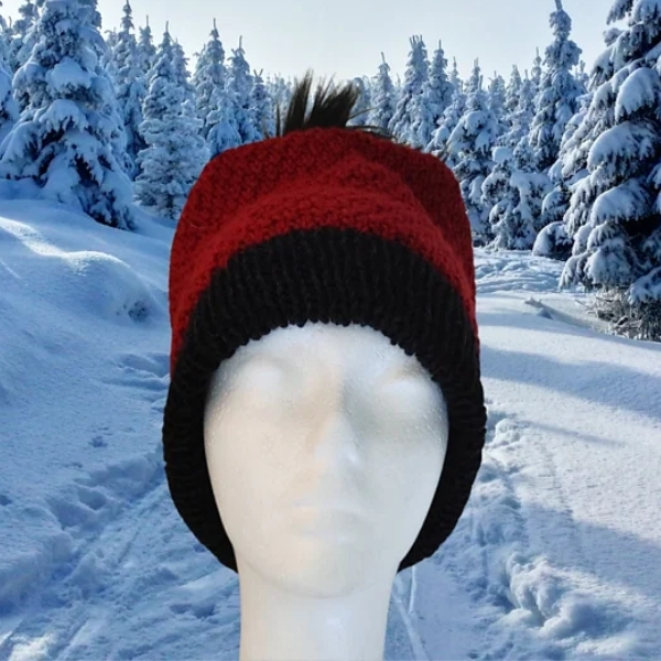 Front view of a red knitted hat with a wide black brim and a black pompom on top, displayed on a white mannequin head in a snowy pine forest.