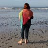 Woman seen from behind, standing barefoot on a pebbly beach and looking out to the waves, wearing a crocheted shawl that shades from coral pink across her shoulders to lime green and turquoise down the point.