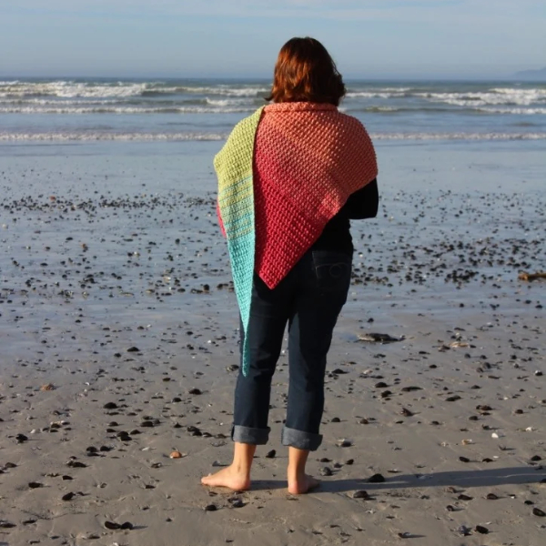 Woman seen from behind, standing barefoot on a pebbly beach and looking out to the waves, wearing a crocheted shawl that shades from coral pink across her shoulders to lime green and turquoise down the point.