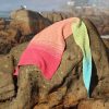 Crocheted shawl laid over a large rock, shading from hot pink through peach to lime green and turquoise at the point, with seagulls perched on rocks in the background.