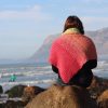 Woman seen from behind, sitting on a rock above the sea and looking towards a mountain, wearing a coral pink crocheted shawl with a lime green collar.