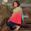 Woman crouching barefoot on a rock and looking to the side, wearing a crocheted shawl that shades from peach at the shoulders to deep coral pink, with a lime green and turquoise end hanging over one shoulder.