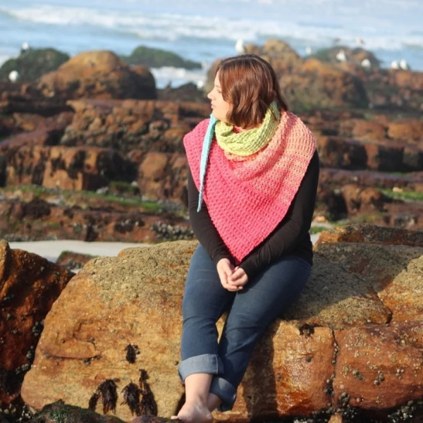 Woman sitting on a rock covered in barnacles and looking to the side, wearing a hot pink crocheted shawl with a lime green cowl and a turquoise tail, with seagulls on the rocks behind her.