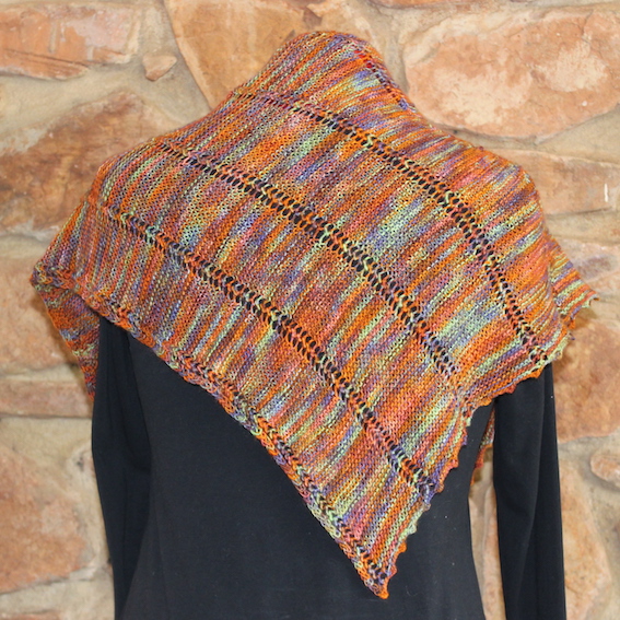 Back view of a hand dyed triangular shawl in orange, purple and green, with dark eyelet lines across the panels and a scalloped edge, on a black mannequin against a stone wall.