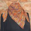 Triangular shawl in orange, purple and green hand dyed yarn, worn gathered at the neck with both points hanging down the front, in front of a stone wall.