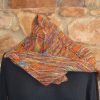 view of a hand dyed shawl in orange, green and purple, fastened with a wooden shawl pin and draped over the shoulders of a black mannequin