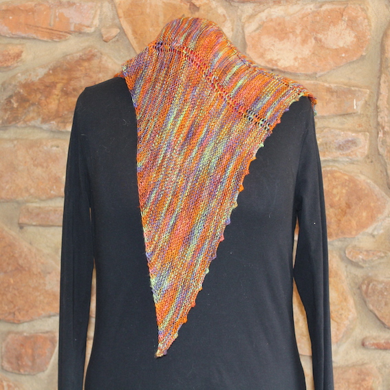 Long point of a hand dyed multicoloured shawl hanging down the front of a black mannequin, showing the striped garter stitch and a scalloped edge