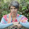 Woman with short grey hair looking at the camera, wearing a crocheted Wild Winds Shawl in bands of coral pink, peach, turquoise, lilac and soft grey, with her arms folded over the back of a chair among green leaves.