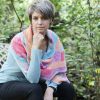Woman with short grey hair sitting with her fist resting at her chin, wearing the Wild Winds Shawl draped over her shoulders, with panels of pink, peach, turquoise, purple and grey crochet and a garden behind her.