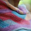 Close up of the Wild Winds Shawl showing an open mesh crochet stitch in bands of red, purple, turquoise, grey and peach.