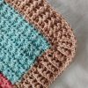 Corner of a crochet blanket showing a turquoise star stitch section edged with a rounded tan ribbed border, resting on soft grey fabric