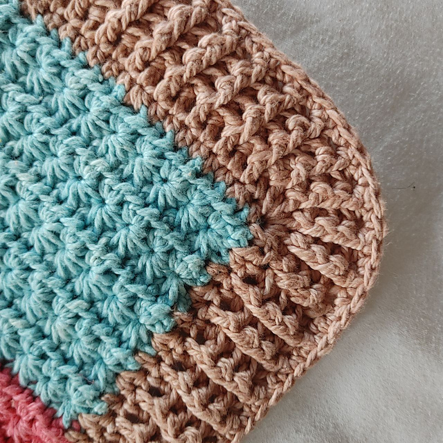 Corner of a crochet blanket showing a turquoise star stitch section edged with a rounded tan ribbed border, resting on soft grey fabric