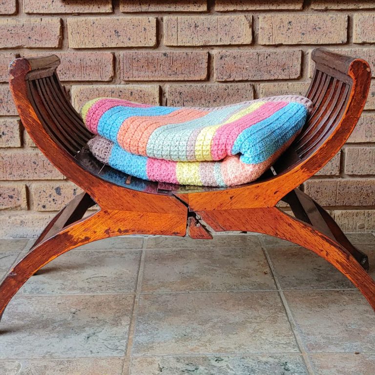 Pastel striped crochet blanket in blue, pink, yellow, peach and sage folded on a curved wooden stool in front of a brick wall
