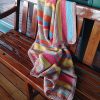 Striped crochet blanket in coral, yellow, aqua, sage and tan draped over the back and seat of a polished wooden bench