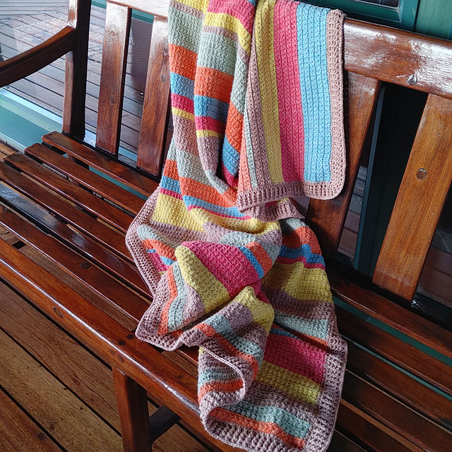 Striped crochet blanket in coral, yellow, aqua, sage and tan draped over the back and seat of a polished wooden bench