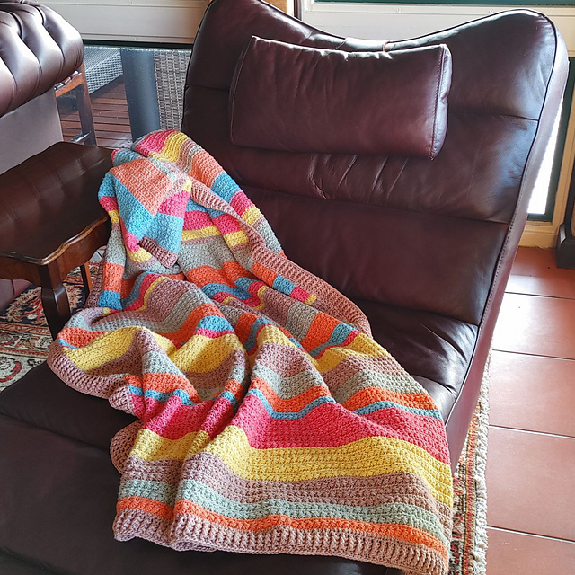 Brightly striped crochet blanket in yellow, pink, orange, sage and blue draped over a brown leather armchair on a terracotta tiled floor.